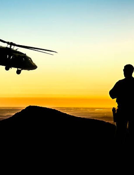 A military helicopter flies over troops on the ground at sunset.